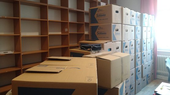 Ein großer Stapel Umzugskartons. Dahinter ein langes, hohes Regal das leer ist.